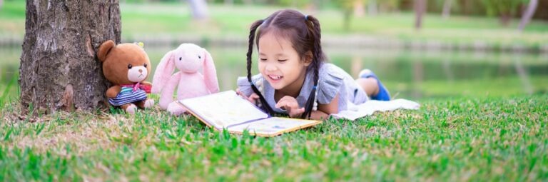 une jeune fille avec des couettes lit un livre dehors avec ses peluches